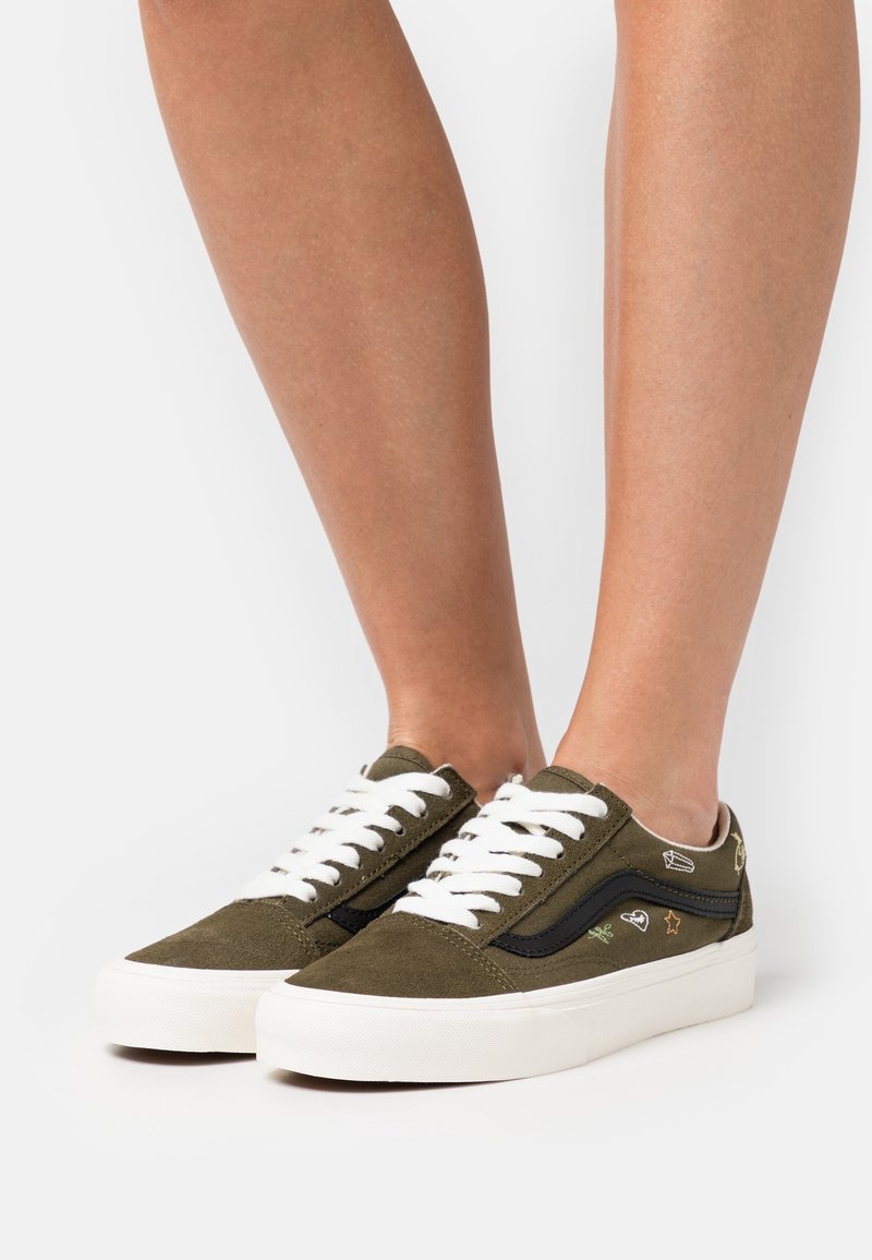 Zapatillas de ante verde oliva con cordones blancos y una franja lateral negra. Presentan detalles bordados decorativos en los lados exteriores. Suelas de goma blanca.