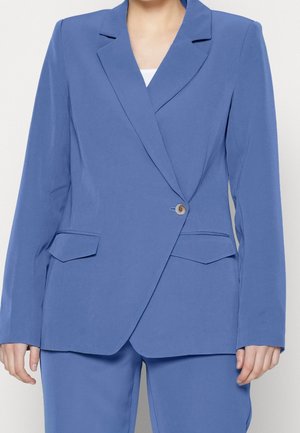 Femme portant un blazer bleu et un pantalon assorti, avec un seul bouton beige et deux poches à rabat sur le blazer.