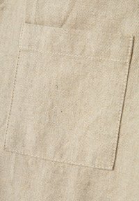Tela beige con una superficie texturizada, que presenta un pequeño bolsillo cuadrado cosido en el material. Diseño simple y utilitario.
