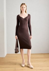 Abito a maglia a coste marrone con maniche lunghe e scollo a cuore, lungo fino al polpaccio, abbinato a tacchi beige e una borsa a spalla.