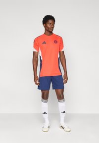 Άνδρας που φορά μπλούζα ποδοσφαίρου πορτοκαλί και μπλε με λογότυπο, μπλε σορτς, λευκές κάλτσες μέχρι τα γόνατα και λευκά παπούτσια ποδοσφαίρου, στέκεται μπροστά σε απλό φόντο.