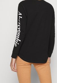 Zwart T-shirt met lange mouwen en witte "Abercrombie" tekst op de linkermouw. Zachte katoenen stof met een ronde zoom. Gecombineerd met geribde tan kleurige broek.