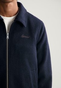 Marine blauwe sweatshirtjack met een textuur, voorzien van een kraag, zijzakken en een geborduurd logo in bruin. Zichtbaar zilveren ritshardware.