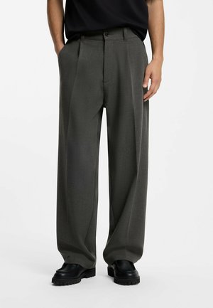 Mann trägt dunkelgraue weite Hose und schwarze Schuhe, steht vor einem schlichten hellen Hintergrund mit entspannten Händen.