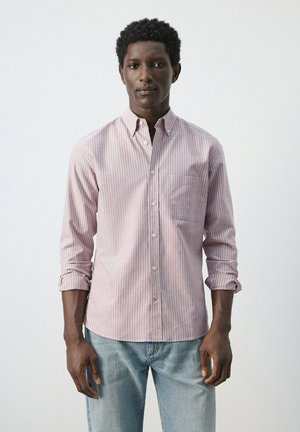 Mann trägt ein rosa-weiß gestreiftes Hemd mit hochgekrempelten Ärmeln und hellblaue Jeans, steht vor einem schlichten Hintergrund.