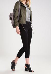 Olivově zelená zipová bunda, pruhovaná halenka, černé slim-fit kalhoty a černé otevřené lodičky na podpatku. Šedý batoh přes rameno.