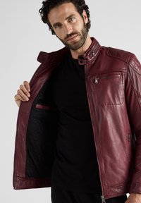 Burgunderfarbene Lederjacke, strukturiertes Design, gestepptes Futter, Reißverschlussfronttasche, silberne Beschläge und hoher Kragen.