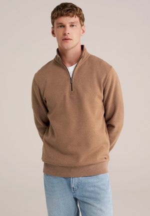 Pullover marrone con zip al collo realizzato in tessuto morbido, con maniche lunghe e polsini a costine, abbinato a jeans azzurri per un look casual.