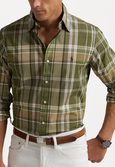 Πράσινη καρό πουκάμισο με κοντό μανίκι, που διαθέτει διακριτές μπεζ και ανοιχτό μπλε ρίγες, φοριέται με καφέ ζώνη και λευκά παντελόνια.