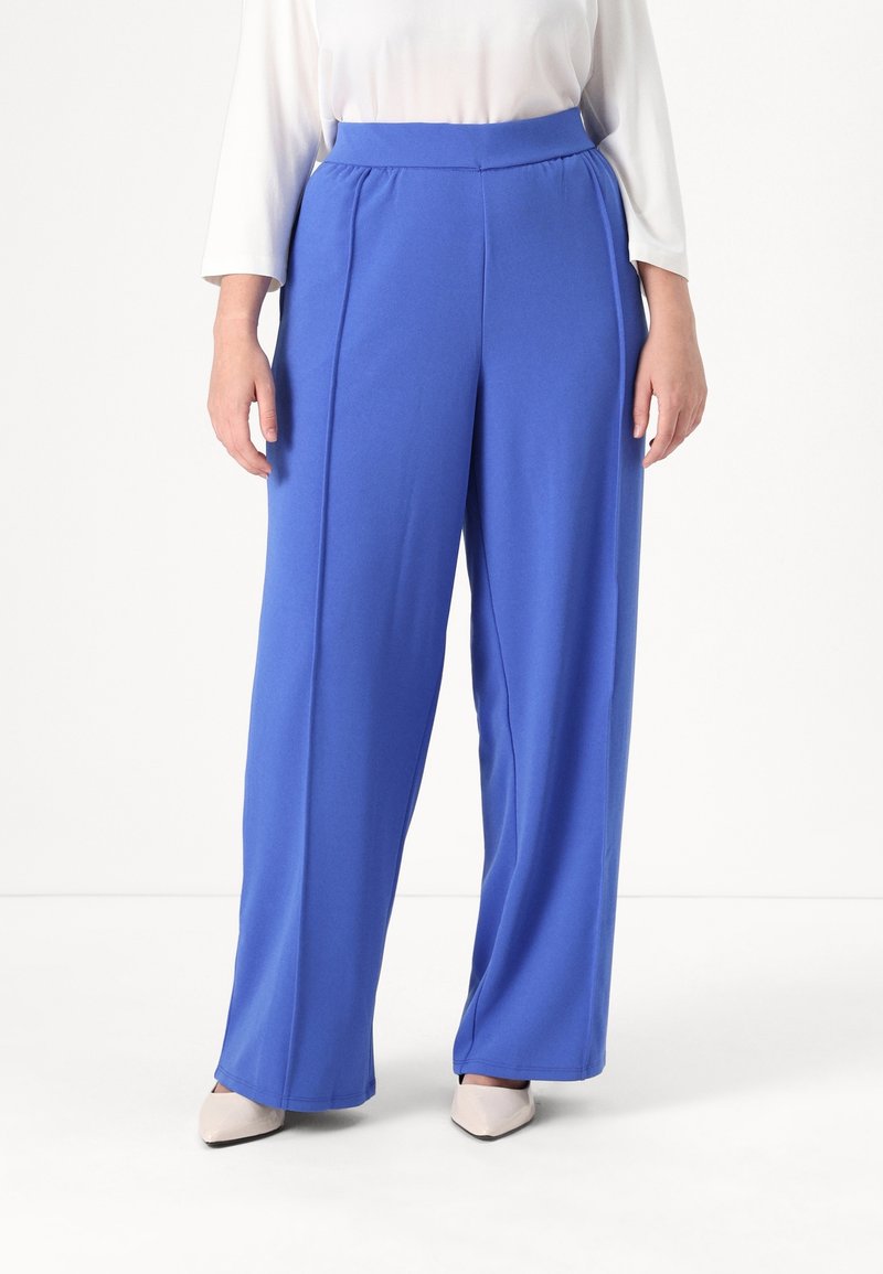 Personne portant un pantalon taille haute à jambes larges bleu royal, associé à des chaussures blanches à bout pointu et un haut blanc à manches longues.