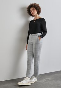 Zwarte blouse met een keyhole-detail, gecombineerd met lichtgrijze, gemêleerde, op maat gemaakte broek. Model draagt casual witte sneakers. Textuurmaterialen en moderne pasvorm.