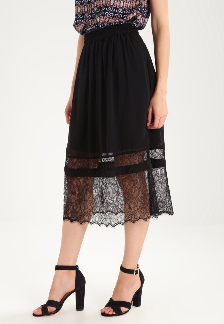 Femme portant une jupe midi noire avec un ourlet en dentelle et des talons hauts à bride autour de la cheville noirs, debout devant un fond blanc uni.