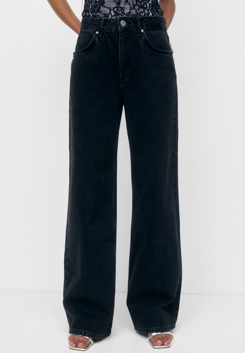 Personne portant un jean noir taille haute à jambes larges, associé à des talons argentés ouverts et un haut en dentelle noire, debout devant un fond uni.