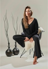 Schwarzes geripptes Langarmtop und weit geschnittene Hose, auf einem Stuhl mit einem Schaffellwurf sitzend. Schwarze Vasen und minimalistische Dekoration im Hintergrund.