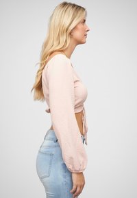 Cropped top rosa chiaro con maniche lunghe e puff, dettagliato con un nodo frontale, abbinato a jeans blu chiaro a vita alta. Texture liscia.