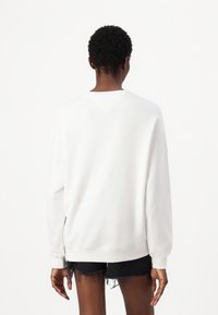 Witte sweatshirt met lange mouwen en geribbelde manchetten, voorzien van een V-vormig detail bij de achterkant van de hals, gecombineerd met zwarte denim shorts.