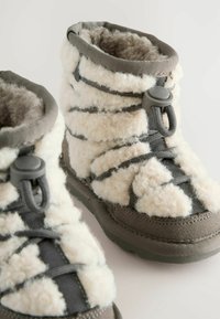 Fuzzy cream and gray boots with a textured outer, adjustable toggle closure, and a soft, insulated lining. Low-cut design for easy wear.