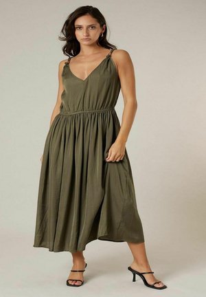 Femme portant une robe midi sans manches vert olive avec taille froncée et sandales à talons noires, debout devant un fond neutre.