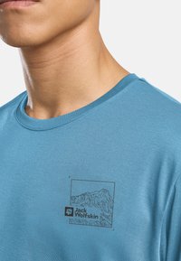 Lichtblauwe katoenen t-shirt met ronde hals en een zwart berggraphic met het "Jack Wolfskin" logo op de linkerkant van de borst.