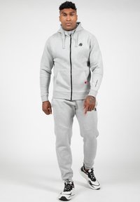 Lichtgrijze zip-up hoodie met trekkoorden, voorzakken en een logo, gecombineerd met bijpassende joggingsbroek, gedragen met zwart-witte sneakers.