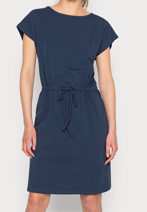 Femme portant une robe décontractée bleu marine avec manches courtes et taille à cordon, debout devant un fond clair uni.