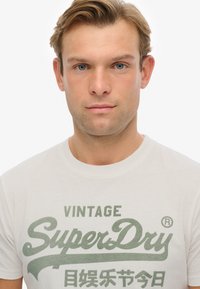 Camiseta de algodón blanca con estampado gráfico en verde "Vintage SuperDry". Diseño de cuello redondo, mangas cortas, tela ligeramente texturizada.