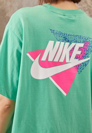 Persoon draagt een muntgroen shirt met een groot wit Nike-logo over roze en blauwe geometrische vormen op de achterkant.