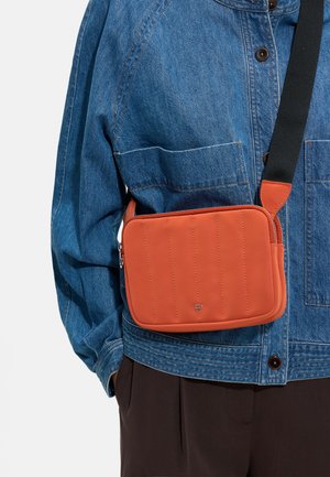 Osoba nosząca niebieską kurtkę dżinsową i ciemne spodnie z pomarańczową torbą przewieszoną przez ramię z czarnym paskiem na klatce piersiowej.