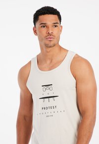 Bežová tielko vyrobené z bavlny, s grafickým dizajnom obsahujúcim "POV" a "PROTEST SPORTSWEAR EST.93" v čiernom tlači. Jednoduchý, priliehavý strih.