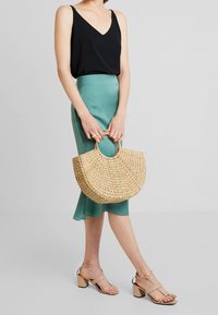 Sac en paille naturelle tressée de forme semi-circulaire, associé à un haut noir, une jupe bleu sarcelle et des sandales à talons carrés beiges.