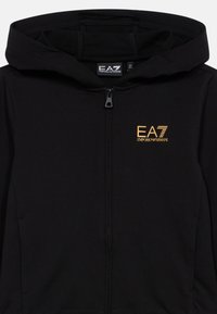 Sudadera con capucha negra con cremallera, hecha de tela suave, con un logo dorado, bolsillos delanteros y capucha con cordón.