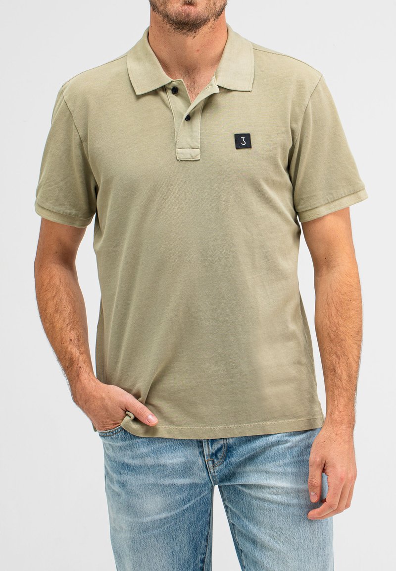Olivgrünes Poloshirt aus strukturiertem Baumwollstoff, mit einer Knopfleiste mit drei Knöpfen, kurzen Ärmeln und einem kleinen schwarzen Logo-Patch auf der Brust.