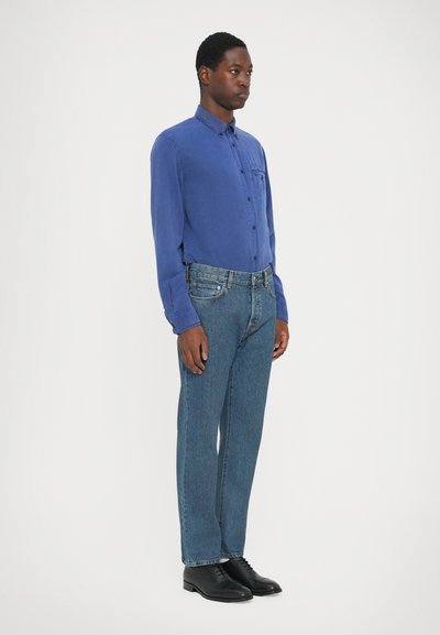 Cămașă albastră cu mâneci lungi, dintr-un material ușor, slim fit, asortată cu blugi drepți de un albastru mediu și pantofi negri cu șireturi.