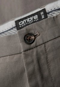 Pantaloni in tessuto grigio in primo piano che presentano una chiusura con bottone, una trama texturizzata e un'etichetta con design in bianco e nero.