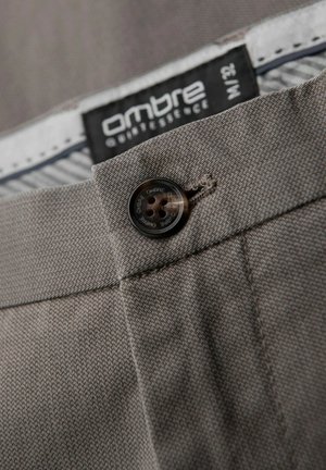 Primer plano de unos pantalones de tejido gris con cierre de botón, tejido texturizado y una etiqueta de marca con diseño en blanco y negro.