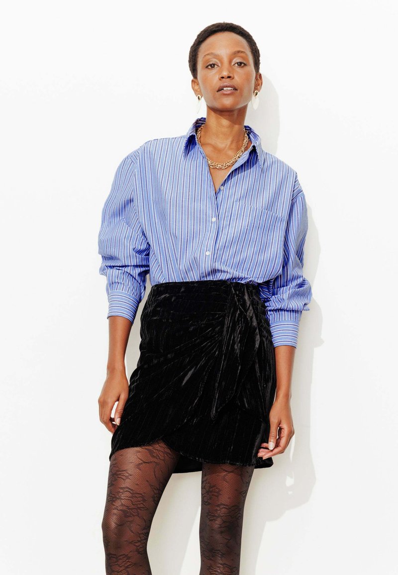 Chemise rayée bleu et blanc avec manches retroussées, associée à une jupe portefeuille en velours noir, et des collants noirs à motifs. Bijoux minimalistes en accent.
