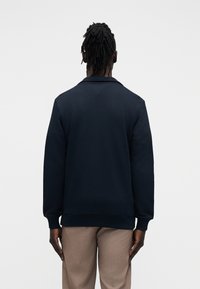 Άνδρας με dreadlocks που φοράει μπλε σκουρόχρωμο πουλόβερ και μπεζ παντελόνια, στέκεται πλάτη σε απλό λευκό φόντο.