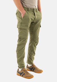 Pantaloni cargo verde oliva con multiple tasche, gamba affusolata e un morbido misto cotone. Abbinati a sneaker marroni con strisce blu.