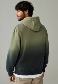 Hoodie s gradijentnim dizajnom, koji prelazi iz svijetlozelene u tamnozelenu. Ima teksturiranu završnu obradu i rebrasti rub.