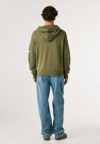 Sudadera de punto verde oliva con un diseño texturizado, que cuenta con capucha y bajo acanalado, combinada con unos vaqueros de mezclilla azul sueltos y zapatos marrones de slip-on.