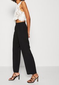 Bílé háčkované crop top spárované s černými, vysokými kalhotami s širokými nohavicemi. Modelka má na sobě černé sandály na vysokém podpatku; minimalistické pozadí.