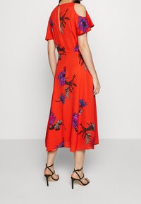 Robe rouge à épaules dénudées avec motif floral violet et vert, coupe fluide, longueur genou, associée à des sandales à talons noirs.