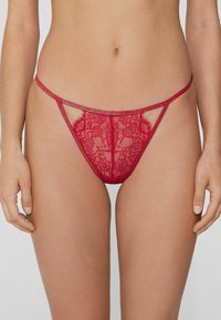 String en dentelle rouge avec tissu transparent, motifs floraux complexes, découpes sur les côtés et fine ceinture.