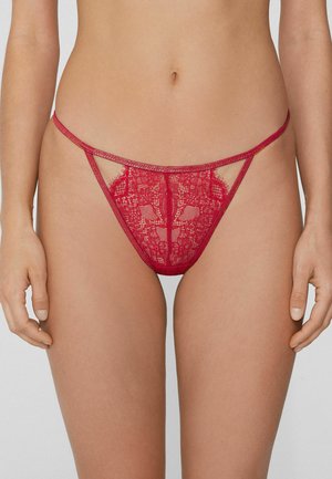 String en dentelle rouge avec tissu transparent, motifs floraux complexes, découpes sur les côtés et fine ceinture.