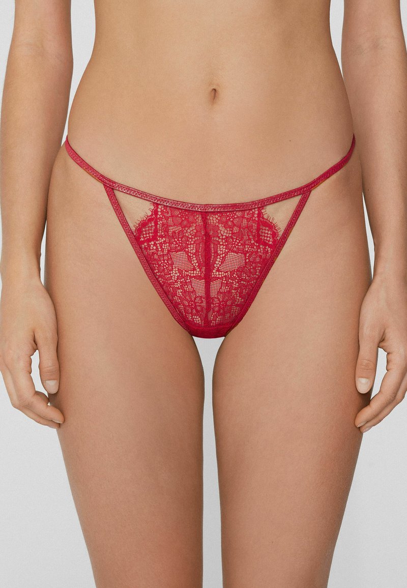 String en dentelle rouge avec tissu transparent, motifs floraux complexes, découpes sur les côtés et fine ceinture.