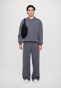 Šedá mikina s dlouhými rukávy a límcem, spárovaná s odpovídajícími tepláky. Černá kabelka na rameno doplňuje outfit. Bílé boty s gumovou podrážkou.