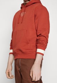 Röd huvtröja i mjukt material, med en ficka framtill, vita randiga muddars och broderad logotyp på bröstet.