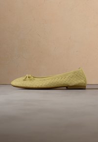 Ballerine tissée jaune avec un petit nœud sur l’avant arrondi, exposée sur une surface grise devant un fond beige.