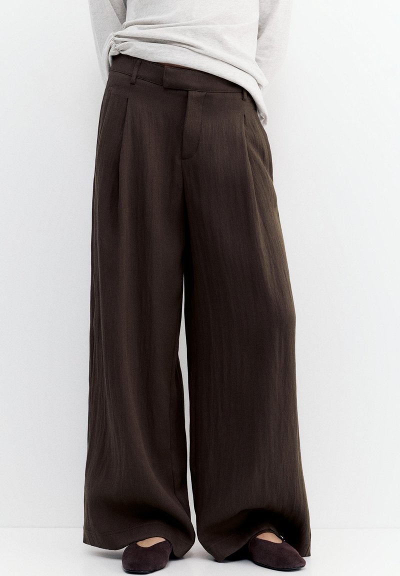 Frau trägt weit geschnittene, dunkelbraune Hosen und schwarze Ballerinas, steht vor weißem, einfarbigem Hintergrund.