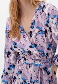 Robe portefeuille à imprimé floral violet avec des manches longues, une taille cintrée avec un lien, et un tissu texturé avec des accents bleus et noirs.